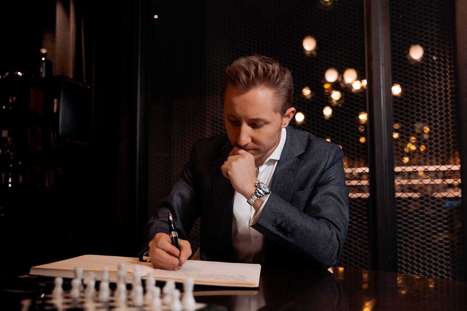Sebastian Klein, Geschäftsführer von Martin&Klein, einer Branding-Agentur aus Salzburg, Österreich, schreibt konzentriert in seinen Block, mit einem Schachbrett im Vordergrund und Lampen im Hintergrund.